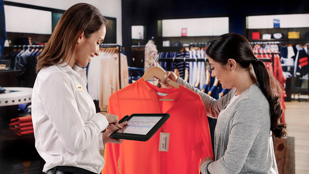 Two women in a store with clothing racks in the background. One of them holding an red shirt and another using a tablet enclosure to check prices.
