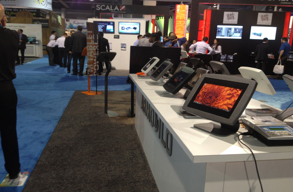 Row of grey and white desktop tablet stands on the Armodilo booth counter at a trade show, beside a card reader