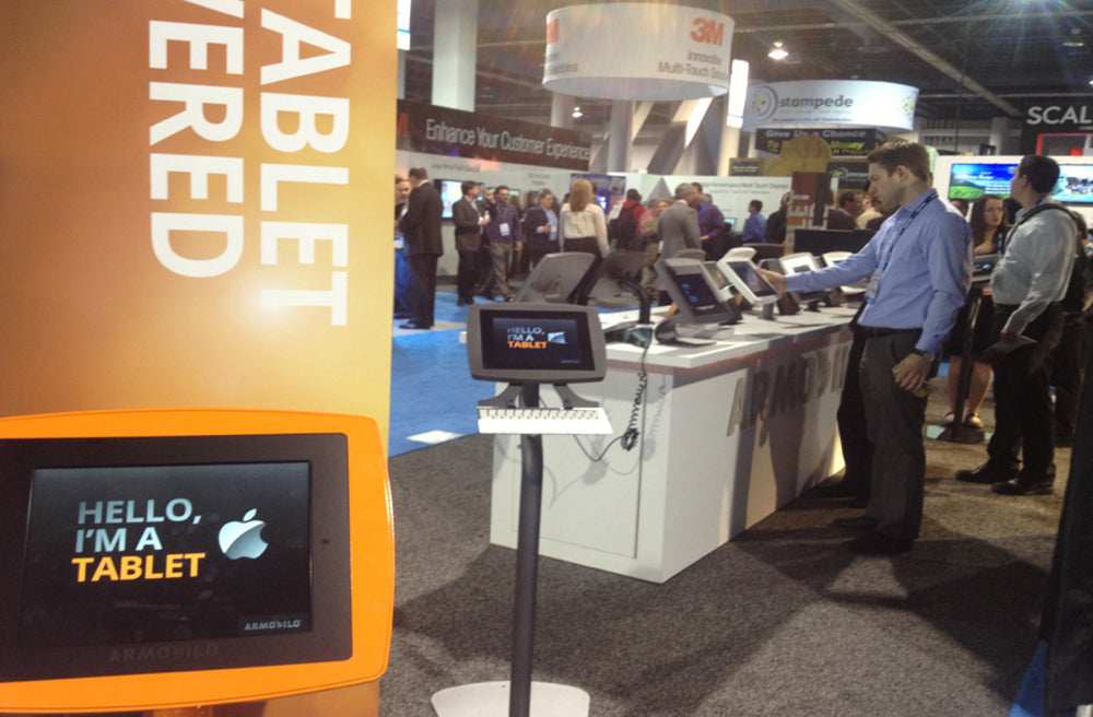 Visitors trying tablet stands at the Armodilo trade show booth, with an orange iPad enclosure in front