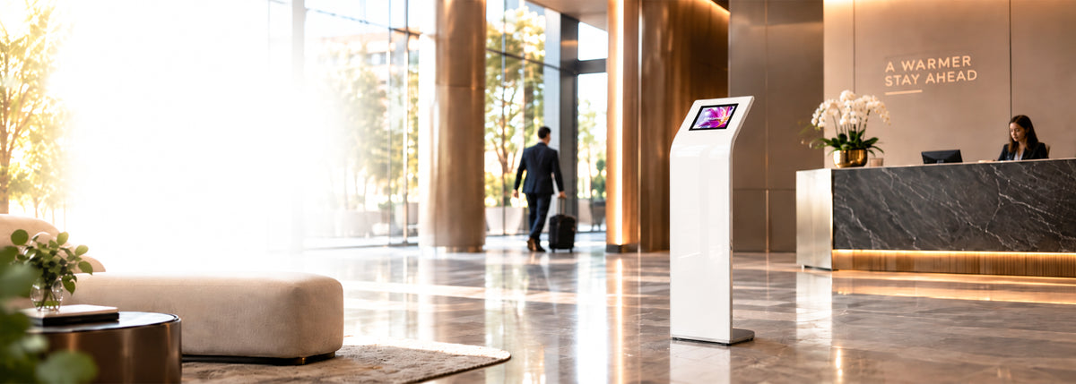 White freestanding tablet kiosk standing in a sunlit hotel lobby beside the reception desk