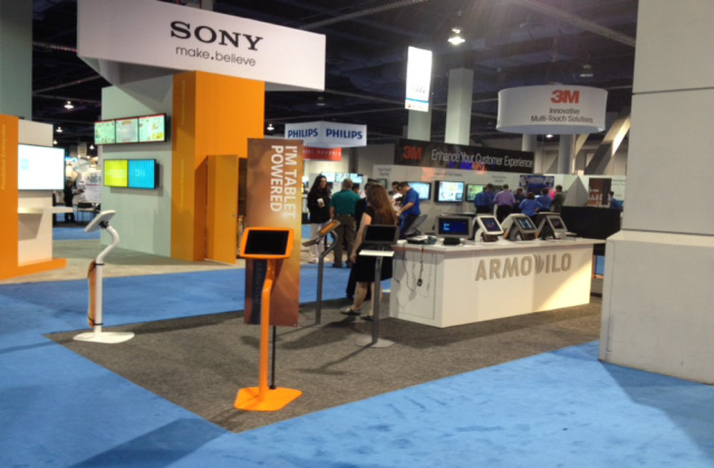 Armodilo trade show booth with orange and white tablet floor stands and a counter of desktop stands